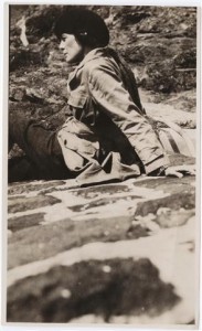 Photograph of H.D. in pants and a jacket leaning back, seated on a blanket on a rocky hill. Undated. From Beinecke Library, Digital Collections, H.D. Papers.