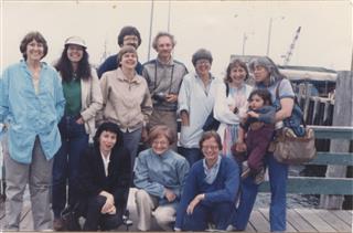 Photo of participants at the HD Centennial Conference in Orono, Maine, 1986. Participants pose for photo on a dock with boats in the background. Pictured from left to right: (front) Anne Friedberg, Charlotte Mandel, Cyrena Pondrom, (back row) Eileen Gregory, Cassandra Laity, (2 participants unidentified), Joe Milicia, Dee Morris, Susan Friedman, Rachel DuPlessis (with child). Photo by Cassandra Laity.